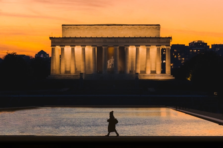 A silhouette of a person walking along the reflecting pool is set against the illuminated Lincoln Memorial at sunset, with a warm orange sky and city buildings in the background.